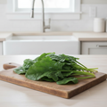 Large broad smooth Summer Solstice spinach leaves on long stems arranged on a rustic wooden board on a bright airy kitchen bench near a silver tap