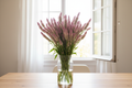 Celosia spicata Flamingo Feather — soft pink-mauve spiky plumes fading to creamy white tips in a clear glass vase in front of a bright window with white sheers slightly open