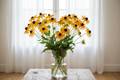 Rudbeckia fulgida Goldsturm black-eyed Susan — tall long-stemmed cut flowers in a clear glass vase, golden-yellow daisy flowers with dark brown-black cones, natural garden-fresh arrangement in front of bright white sheer curtains