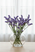 Limonium sinuatum Sky Blue statice, 35 stems in a wide-opening globe fishbowl glass vase, soft lavender-blue-purple periwinkle blooms with elongated open corymbs and characteristic winged stems, loose sheer curtains behind